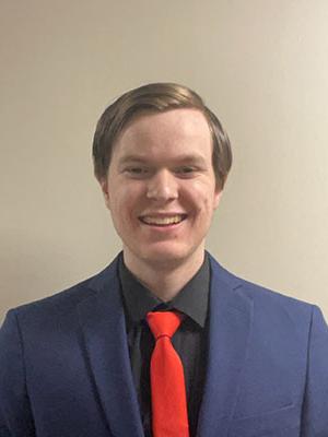 White man with light brown hair wearing a black dress shirt and navy blue suit jacket with a red tie